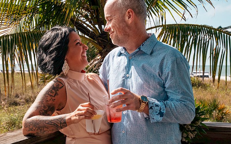 A couple is standing on a wooden deck, smiling at each other and holding drinks, with a palm tree and greenery in the background.