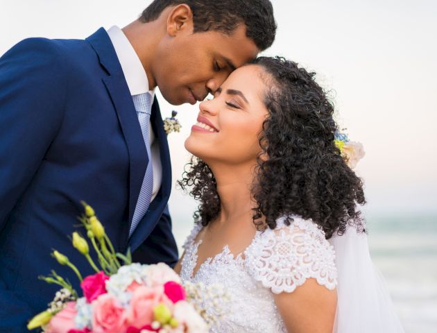 A couple in wedding attire shares a tender moment on a beach, with the groom in a navy suit and the bride holding a bouquet of pink and white flowers.