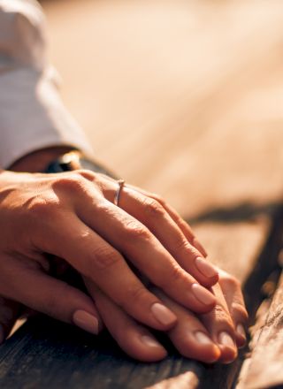 Two hands, one with a wedding ring, rest gently on a wooden surface, suggesting a moment of connection and intimacy.