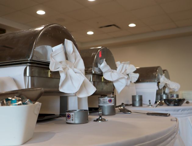 A buffet setup with covered chafing dishes, adorned with white cloth napkins, and a condiment station in a well-lit room.