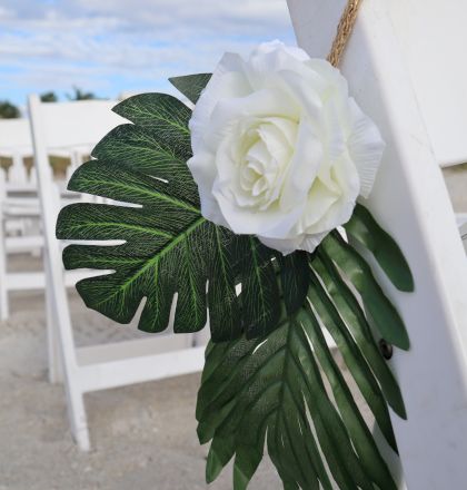 A white rose and green leaves are attached to a white chair, set up outdoors with additional chairs in the background and a cloudy sky above.