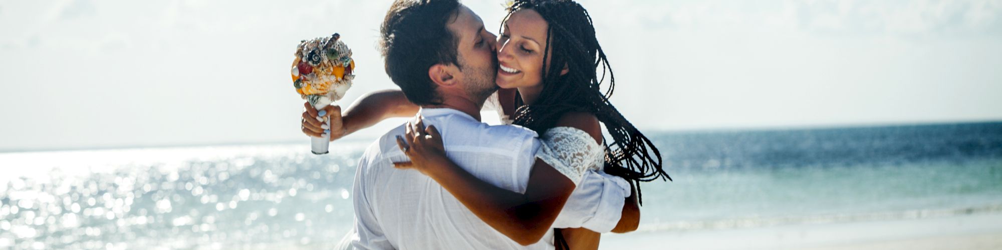 A couple on a beach, with the man lifting the woman who holds a bouquet, both dressed in white, smiling and surrounded by a beautiful coastline.