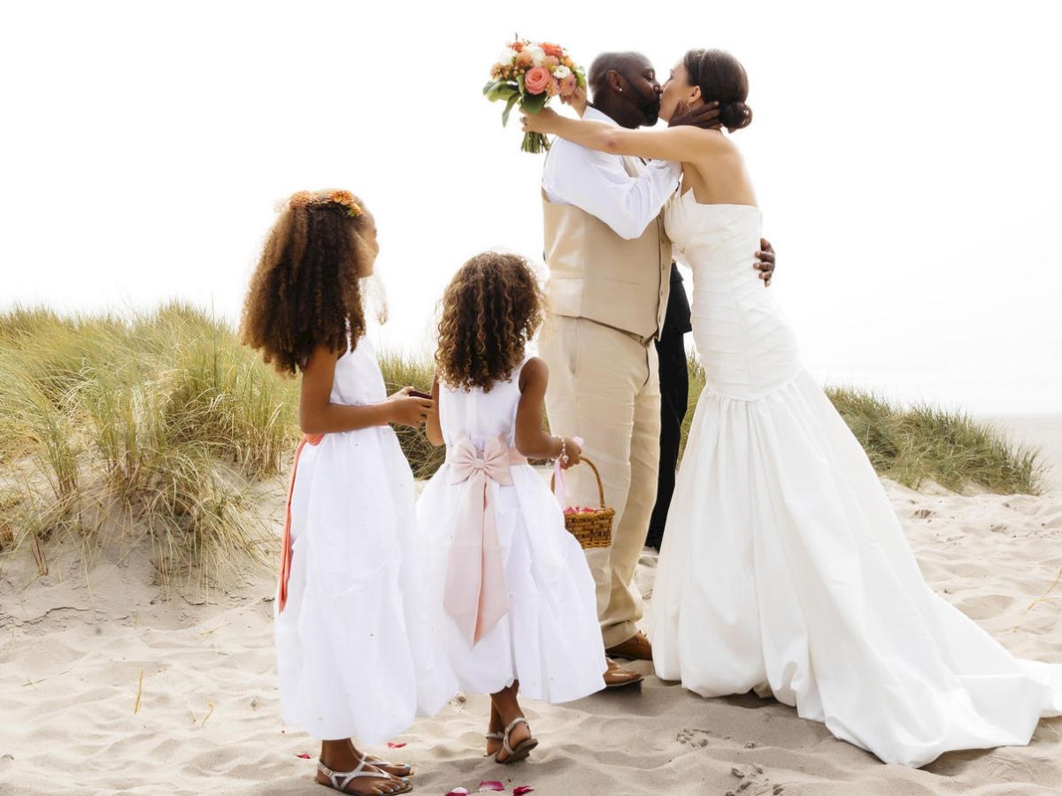 A couple is kissing on a beach, with two flower girls dressed in white standing nearby, holding a flower basket.