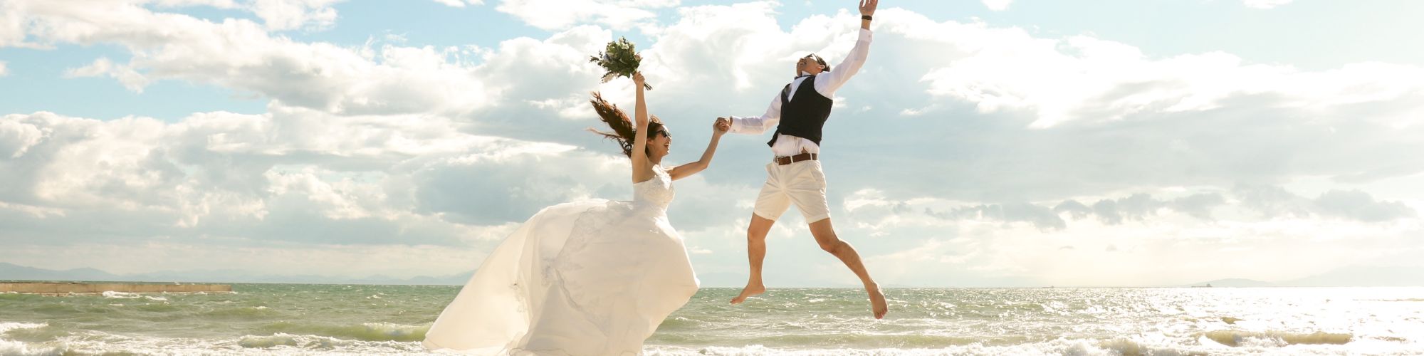 A bride and groom are joyfully jumping on a beach, with the ocean and a partly cloudy sky in the background. They appear to be celebrating.