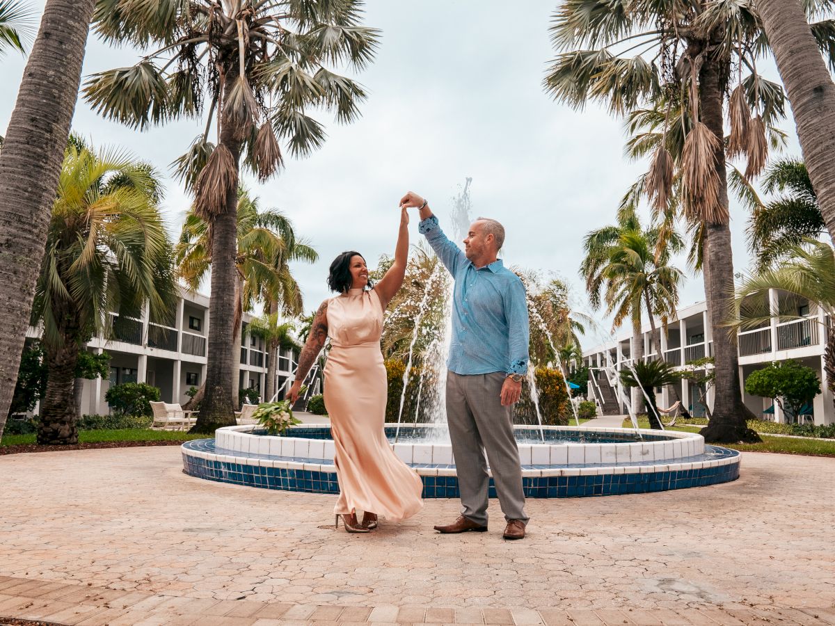 A couple is dancing in front of a fountain, surrounded by palm trees, in what appears to be a resort or courtyard setting.