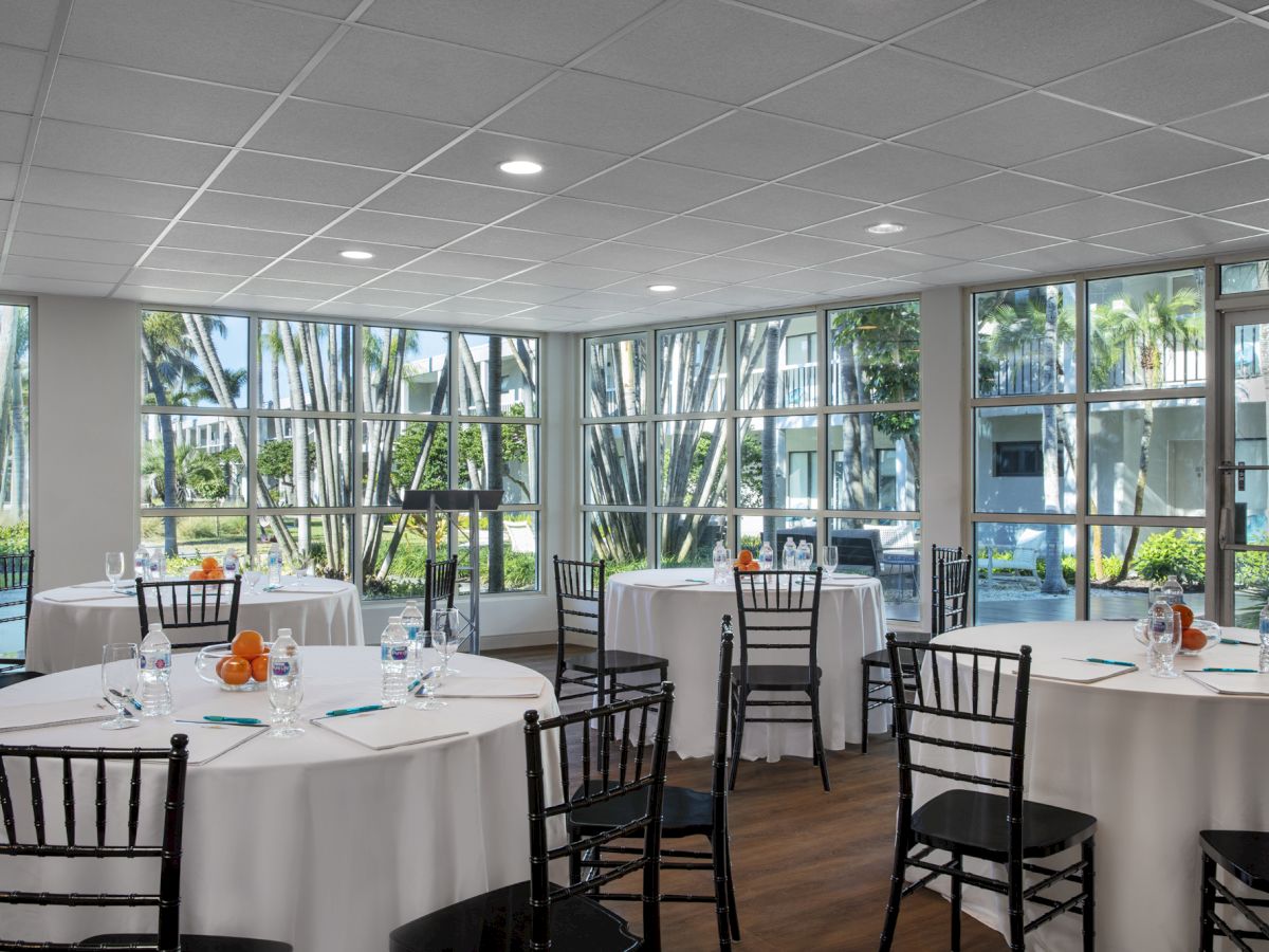 The image shows a bright conference room with large windows, round tables with white tablecloths, black chairs, and notepads on the tables.