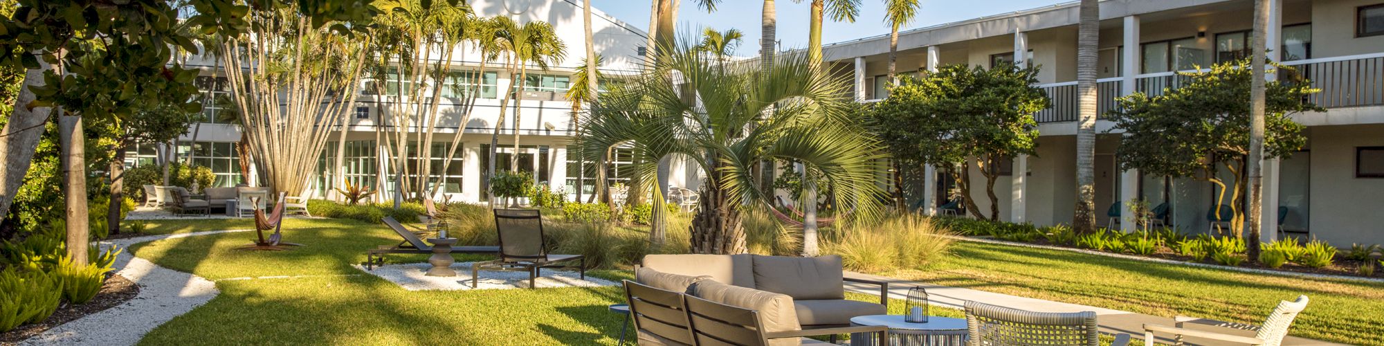 The image shows an outdoor seating area with modern furniture on a patio, surrounded by lush greenery, palm trees, and a building in the background.