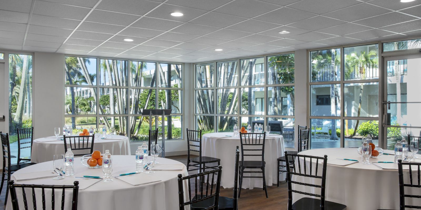 A meeting room with round tables covered in white cloths, black chairs, notepads, water bottles, and fruit. Large windows offer a garden view.