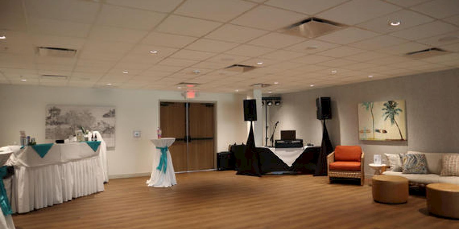 A room set up for an event with tables, chairs, a DJ area with speakers, and decorations including a painting and fabric accents.