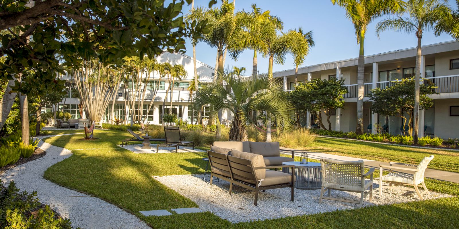 A landscaped courtyard with outdoor seating, palm trees, and a path along the green lawn in front of a two-story building with balconies.