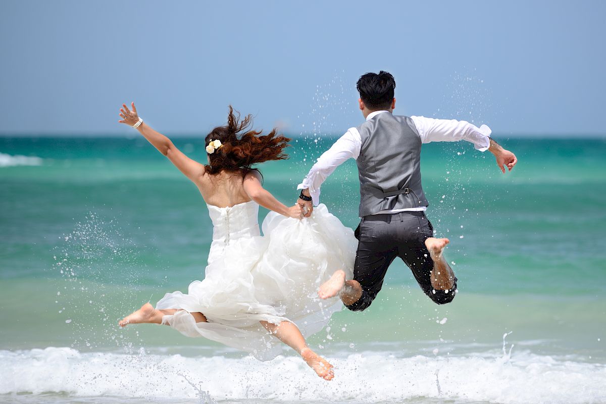 A couple dressed in wedding attire is joyfully jumping into the ocean, holding hands, with the turquoise sea and blue sky in the background.