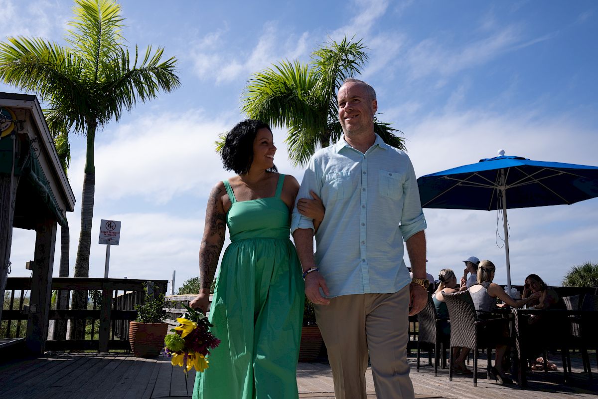 A smiling couple walks arm in arm outdoors near a dining area with palm trees and clear skies in the background, carrying a bouquet of flowers.