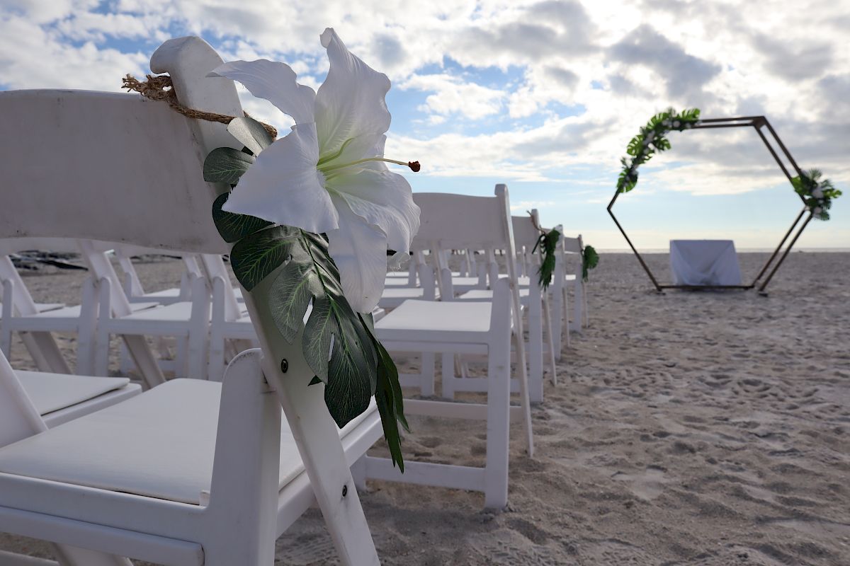 This image depicts a beach wedding setup with white chairs decorated with flowers, and an arch adorned with greenery, under a partly cloudy sky.