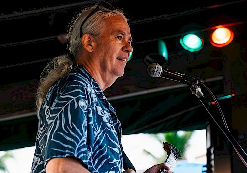 A man with long hair plays an electric guitar and sings into a microphone on stage. He is wearing a patterned shirt under colored lights.