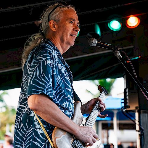 A man with long hair plays an electric guitar and sings into a microphone on stage. He is wearing a patterned shirt under colored lights.