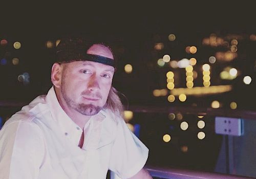 A man with a beard is sitting on a balcony at night with city lights in the background, wearing a white shirt and a black cap.