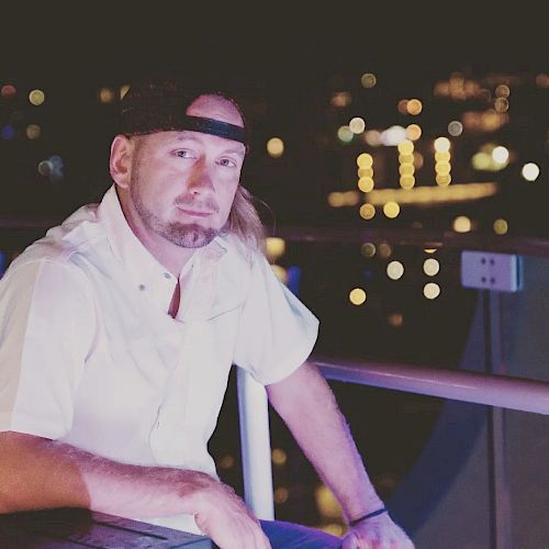 A man with a beard is sitting on a balcony at night with city lights in the background, wearing a white shirt and a black cap.