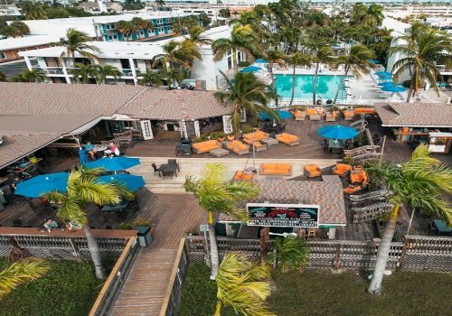 An aerial view of a resort featuring a pool, outdoor seating with umbrellas, palm trees, and adjacent buildings with a tropical atmosphere.