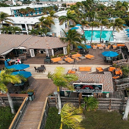 An aerial view of a resort featuring a pool, outdoor seating with umbrellas, palm trees, and adjacent buildings with a tropical atmosphere.