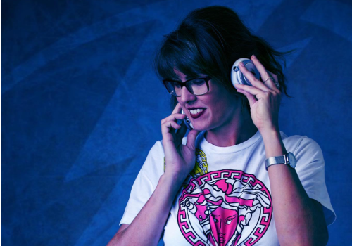 A woman with glasses in a white t-shirt and white pants is on a phone call while also holding headphones against their ear, standing against a blue background.