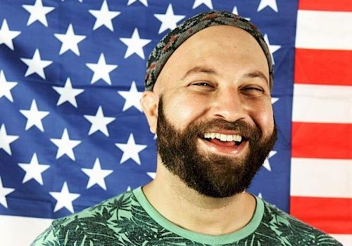 A bearded man with a headband is smiling in front of a U.S. flag, wearing a green shirt with a leaf pattern.