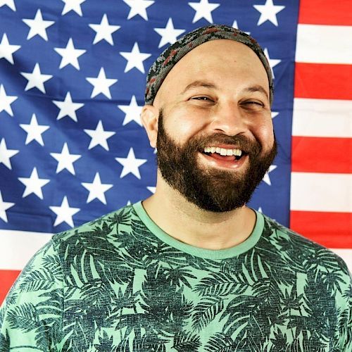 A bearded man with a headband is smiling in front of a U.S. flag, wearing a green shirt with a leaf pattern.