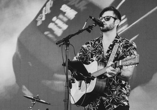 A man is performing on stage with a guitar, singing into a microphone. A drum set and other instruments are in the background, alongside a large screen.