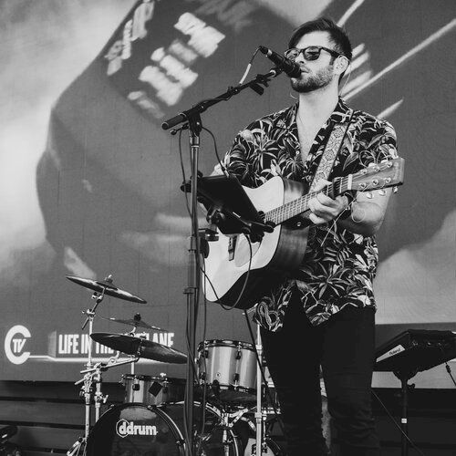 A man is performing on stage with a guitar, singing into a microphone. A drum set and other instruments are in the background, alongside a large screen.