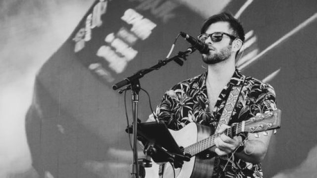 A man is performing on stage with a guitar, singing into a microphone. A drum set and other instruments are in the background, alongside a large screen.