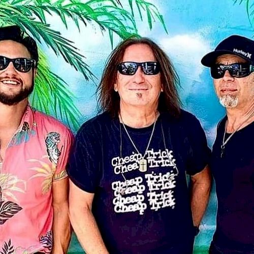 Three men wearing sunglasses, standing in front of a tropical background with palm leaves. One wears a band t-shirt, another a Hawaiian shirt.