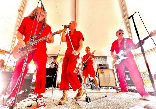 A musical band, dressed in matching red outfits, is energetically performing on stage under a white tent with instruments like guitars and a microphone.