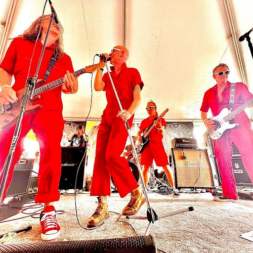 A musical band, dressed in matching red outfits, is energetically performing on stage under a white tent with instruments like guitars and a microphone.