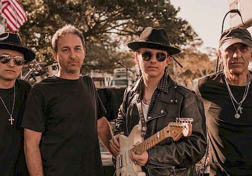 Four men posing outdoors, one holding a guitar; they are dressed in various styles of black clothing and hats.