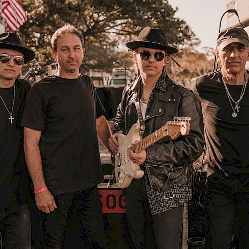 Four men posing outdoors, one holding a guitar; they are dressed in various styles of black clothing and hats.