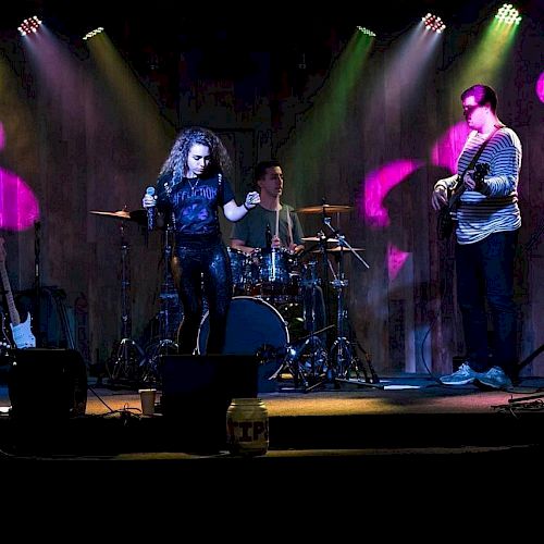 A band performing on stage with colorful lights; the singer is in the center and the drummer and guitarist are to her right and left, respectively.