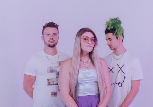 The image shows three people posing against a plain background, with the central person wearing purple pants and sunglasses, and the person on the right with green hair.