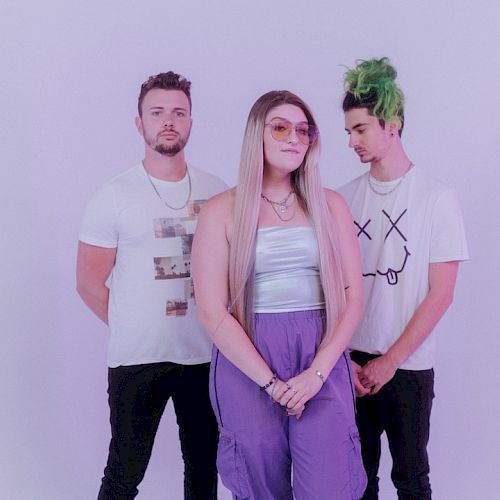 The image shows three people posing against a plain background, with the central person wearing purple pants and sunglasses, and the person on the right with green hair.