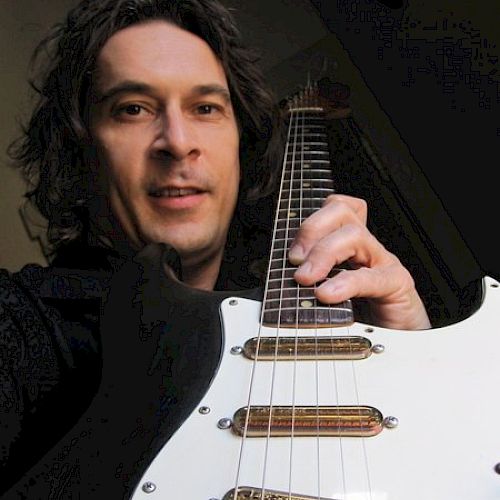 A man holding a white electric guitar, looking at the camera, with dark hair and wearing dark clothing, taking a close-up selfie.