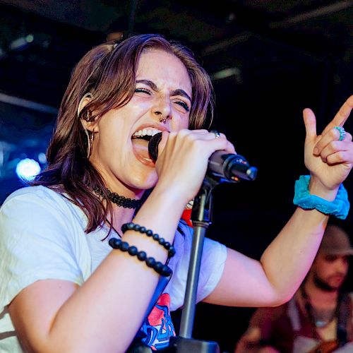 A person passionately singing into a microphone on stage, wearing bracelets and a blue hair tie, with a band performing in the background.