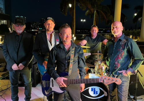 A group of five men, likely a band, pose with musical instruments outdoors at night; drums, bass, and electric guitar are visible.