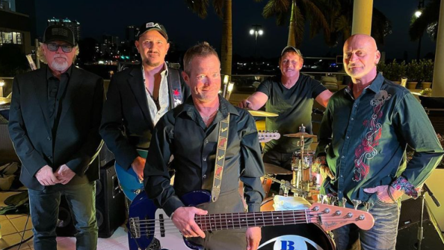 A group of five men, likely a band, pose with musical instruments outdoors at night; drums, bass, and electric guitar are visible.