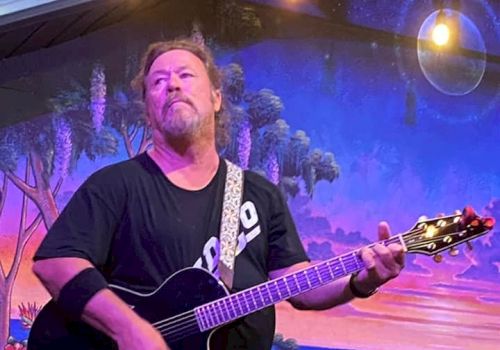 A person is playing an acoustic guitar on stage with a colorful backdrop of a beach sunset scene and hanging lights above them.