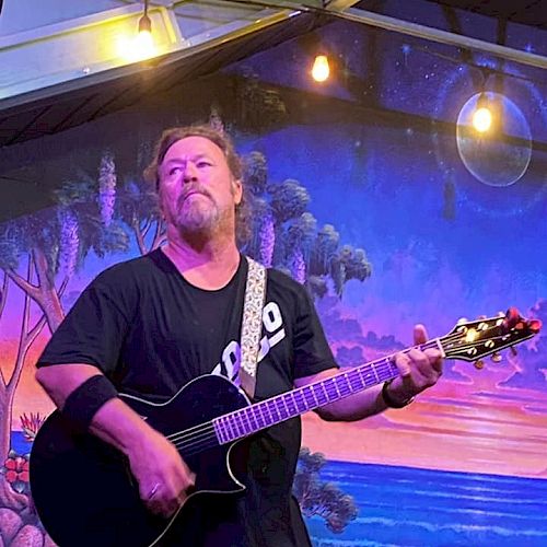 A person is playing an acoustic guitar on stage with a colorful backdrop of a beach sunset scene and hanging lights above them.