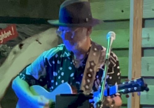 A man wearing a hat and patterned shirt plays an acoustic guitar while standing in front of a microphone, in a dimly lit setting.