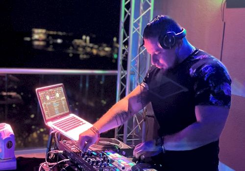 A DJ is performing at a night event, using turntables and a laptop, illuminated by purple and blue lights.