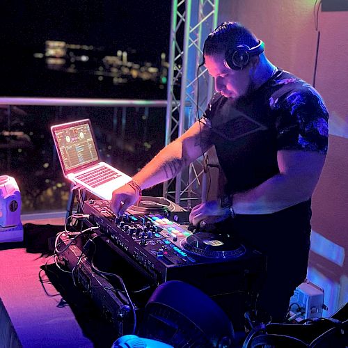 A DJ is performing at a night event, using turntables and a laptop, illuminated by purple and blue lights.