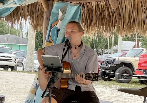 A person is sitting on a stool under a thatched roof, playing a guitar and singing into a microphone, with cars parked in the background.