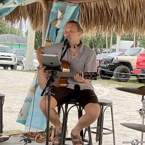 A person is sitting on a stool under a thatched roof, playing a guitar and singing into a microphone, with cars parked in the background.