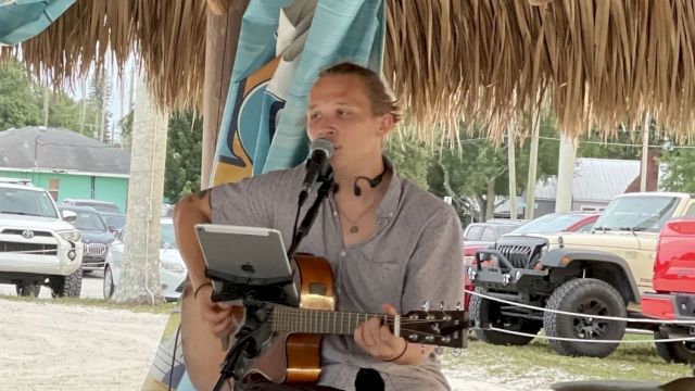 A person is sitting on a stool under a thatched roof, playing a guitar and singing into a microphone, with cars parked in the background.