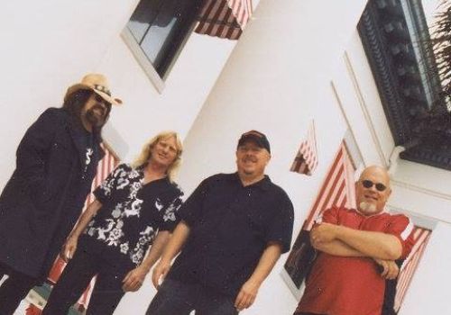 Four men are standing in front of a white building with red-and-white striped awnings. They are posing for the photo outdoors.